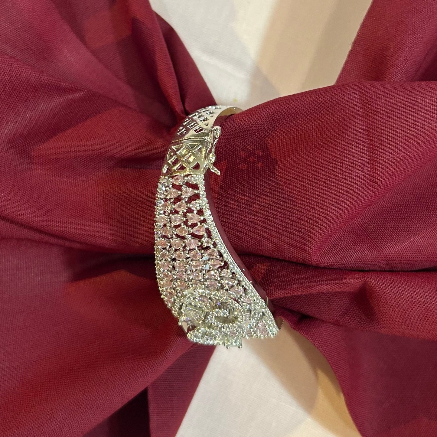 A close-up of a silver-toned, diamond-embellished ring-shaped decorative pin or brooch fastened to a draped burgundy fabric against a light cream background.