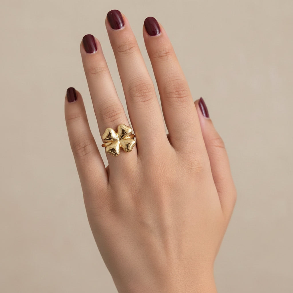 A close-up of a hand wearing a large, textured gold ring with a floral or clover-like design, resting on a dark, curved surface.
