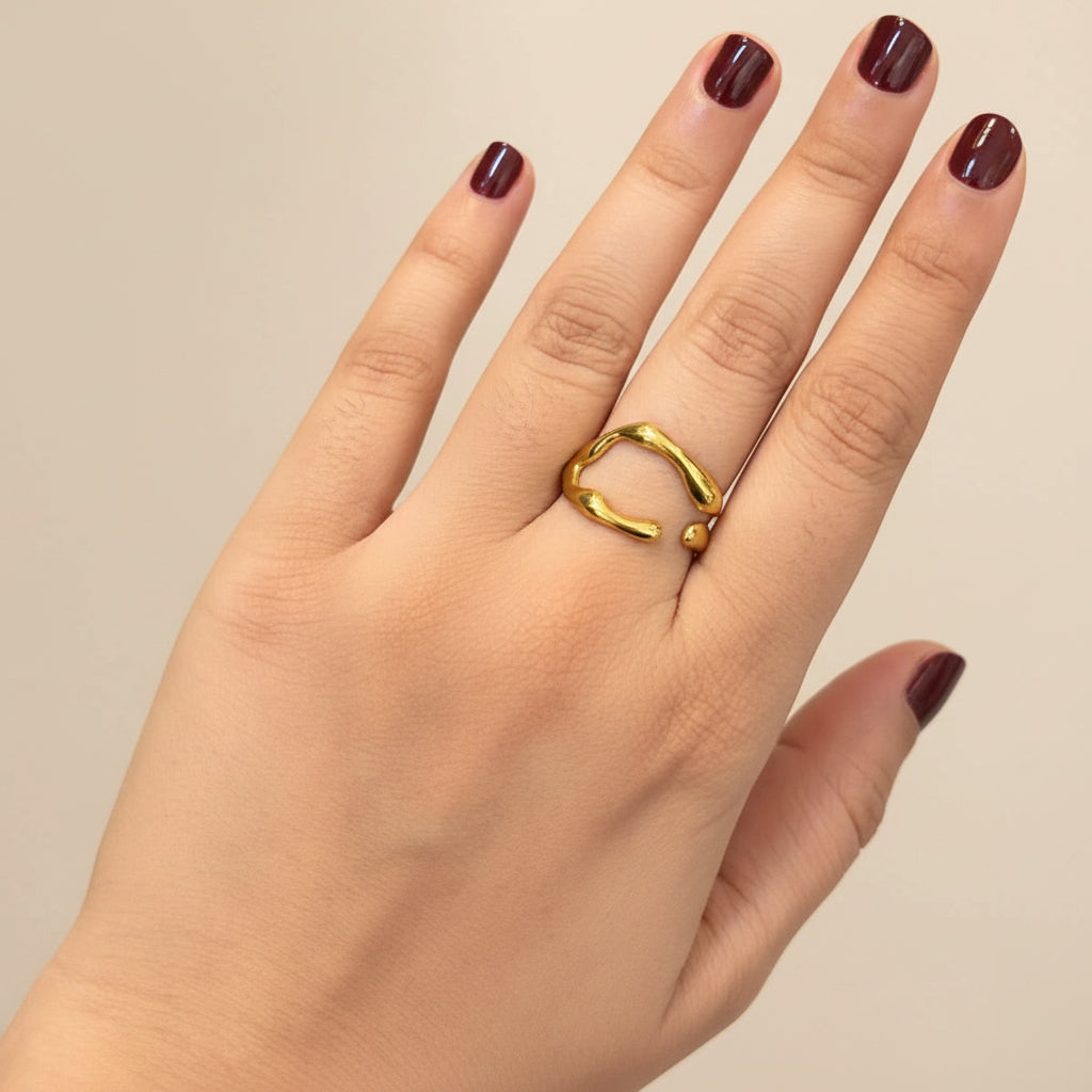 A gold ring with a textured, organic wavy band worn on a person's ring finger, shown against a dark textured surface.
