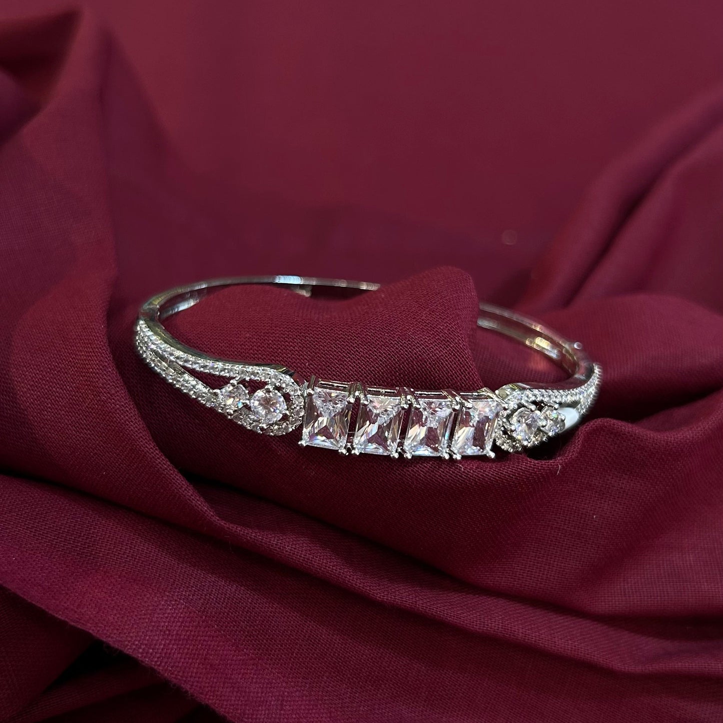 A silver-toned bangle bracelet with a row of clear rectangular and round stones, displayed on draped burgundy fabric.