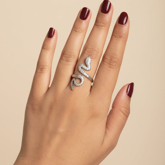 A silver snake ring with small clear stones, worn on a person's ring finger, shown against a black leather surface.