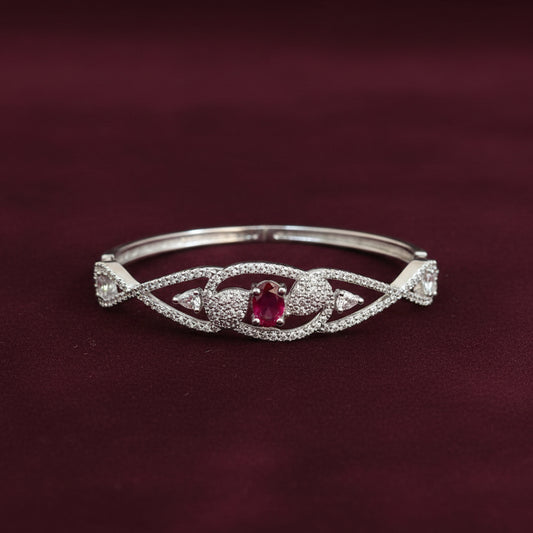 A silver-toned bangle bracelet with a central red gemstone and pavé-set details, displayed on draped burgundy fabric.