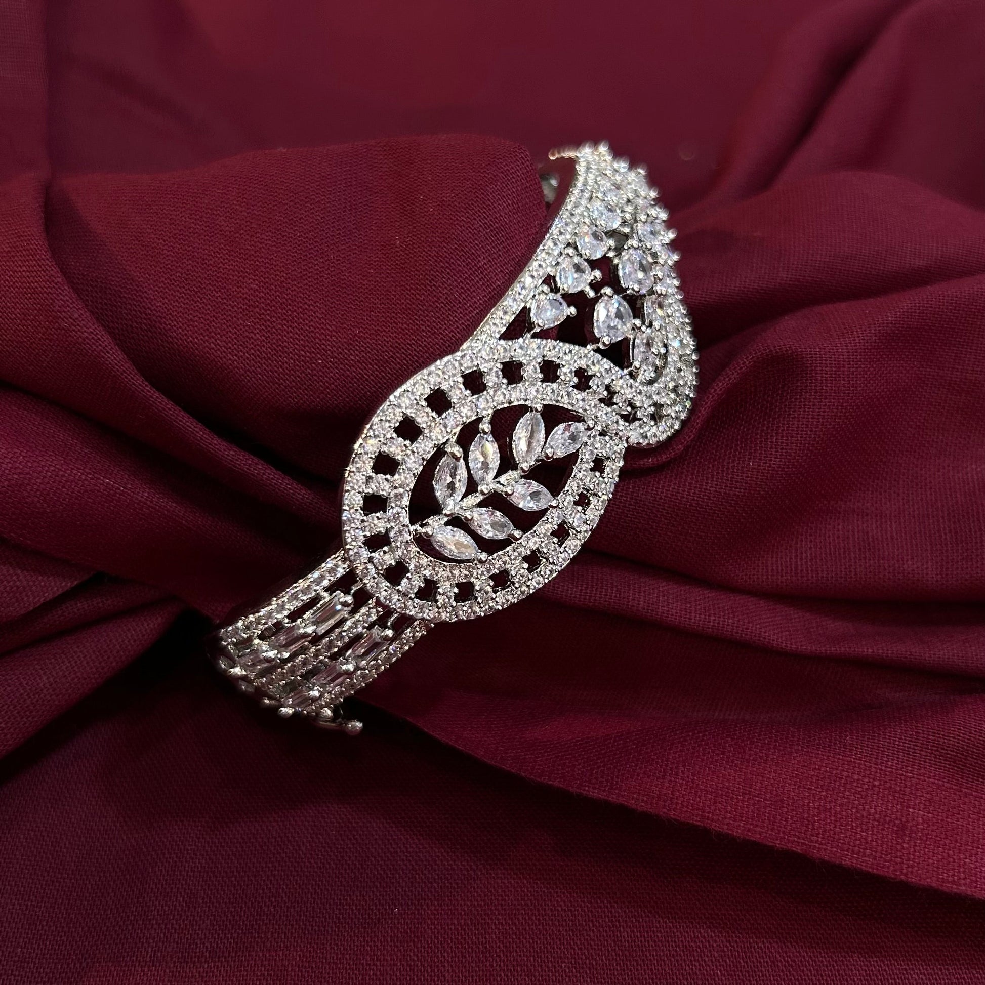 An ornate silver-toned diamond-encrusted cuff bracelet with a leaf motif, displayed on draped burgundy fabric.