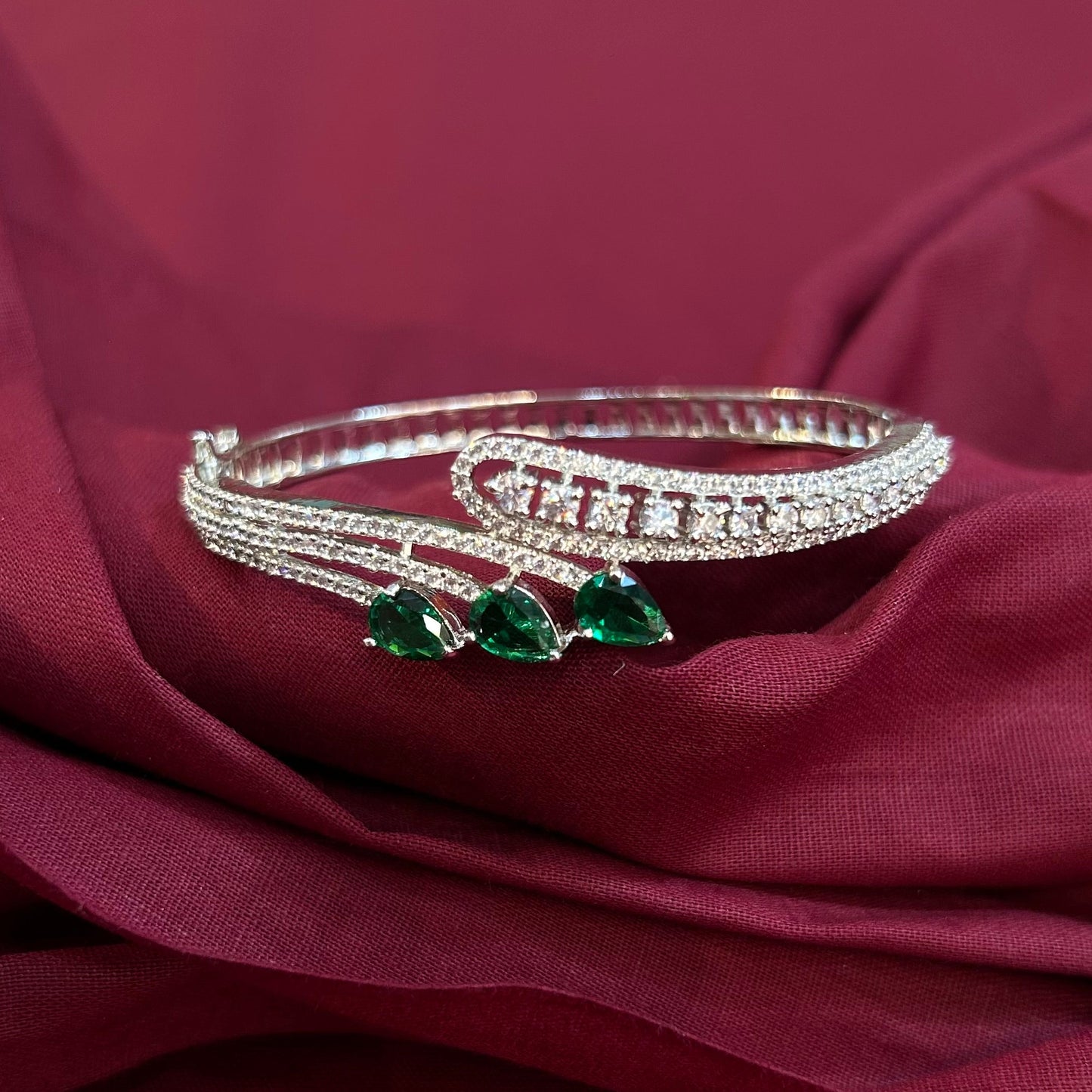 A silver-toned multi-strand bangle bracelet with three green teardrop-shaped gemstones, displayed on draped burgundy fabric.