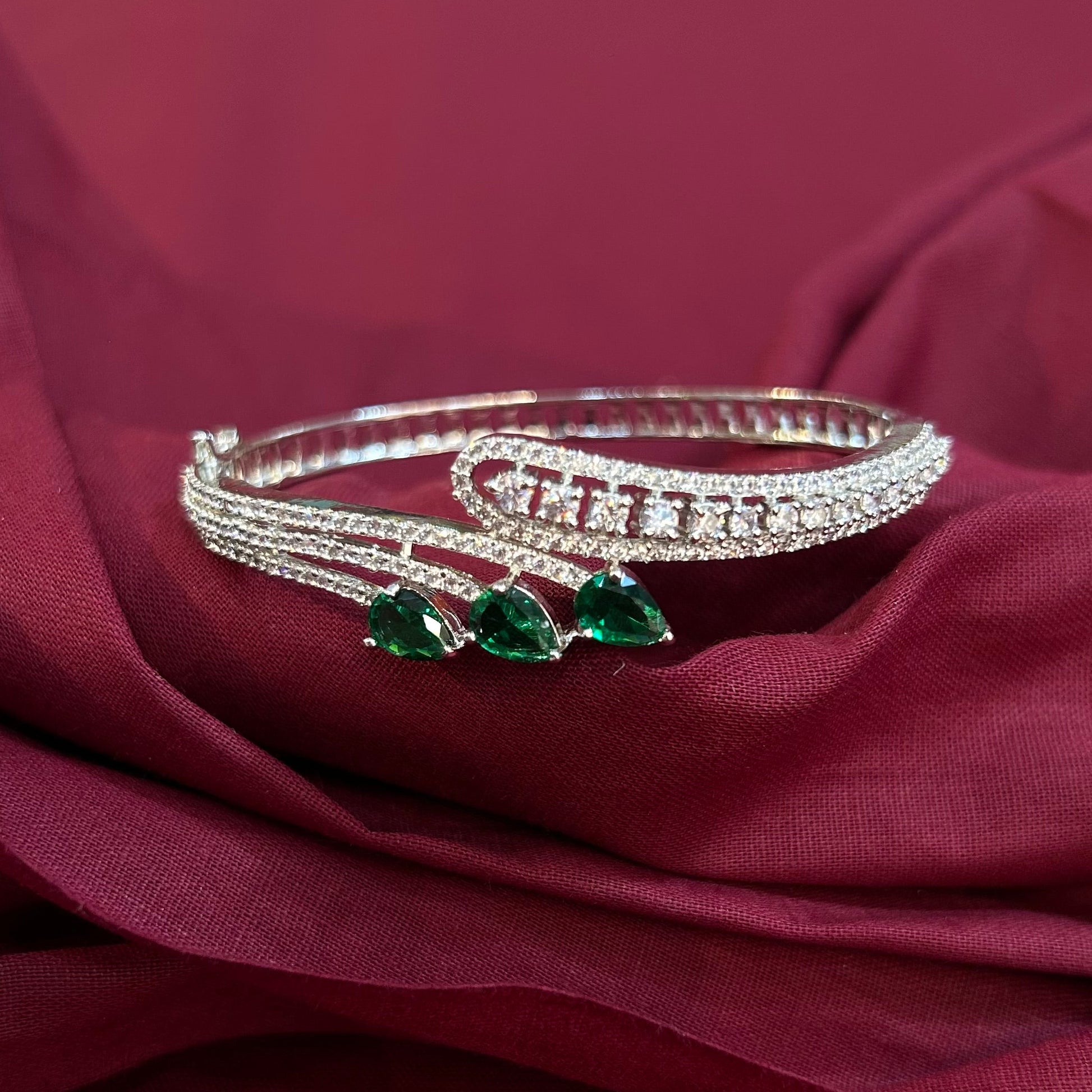 A silver-toned multi-strand bangle bracelet with three green teardrop-shaped gemstones, displayed on draped burgundy fabric.