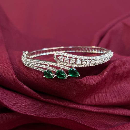 A silver-toned multi-strand bangle bracelet with three green teardrop-shaped gemstones, displayed on draped burgundy fabric.