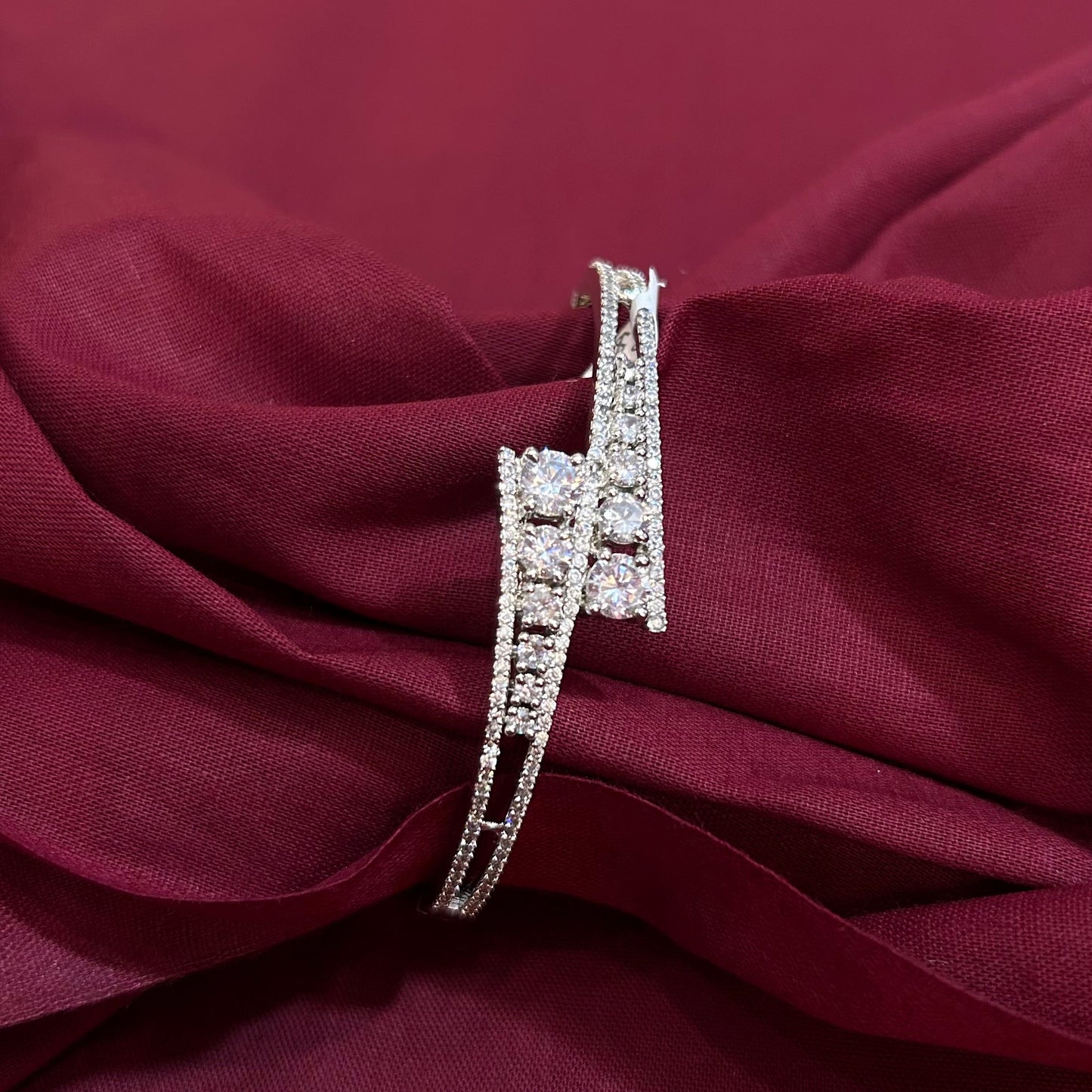 A silver-toned bangle bracelet with pavé-set clear stones, displayed on draped burgundy fabric.