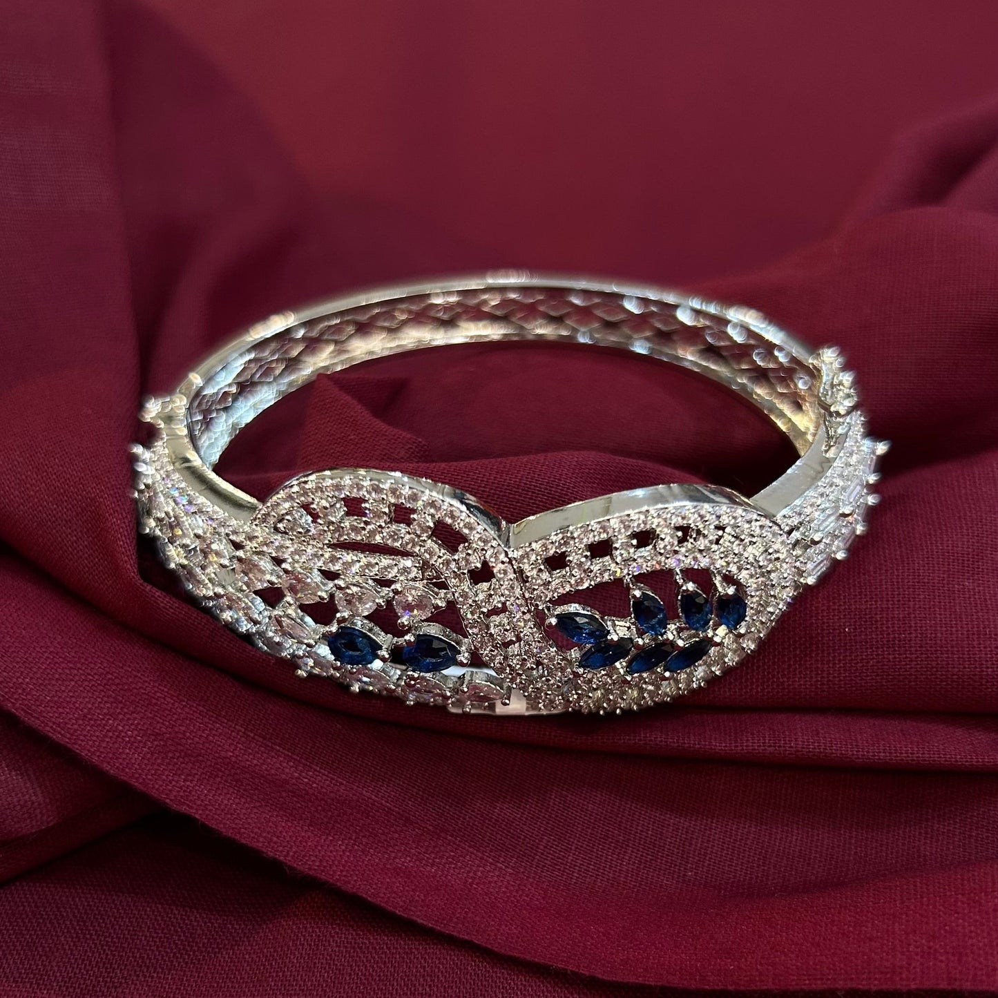 A silver-toned filigree bangle bracelet with dark blue gemstone accents, displayed on draped burgundy fabric.