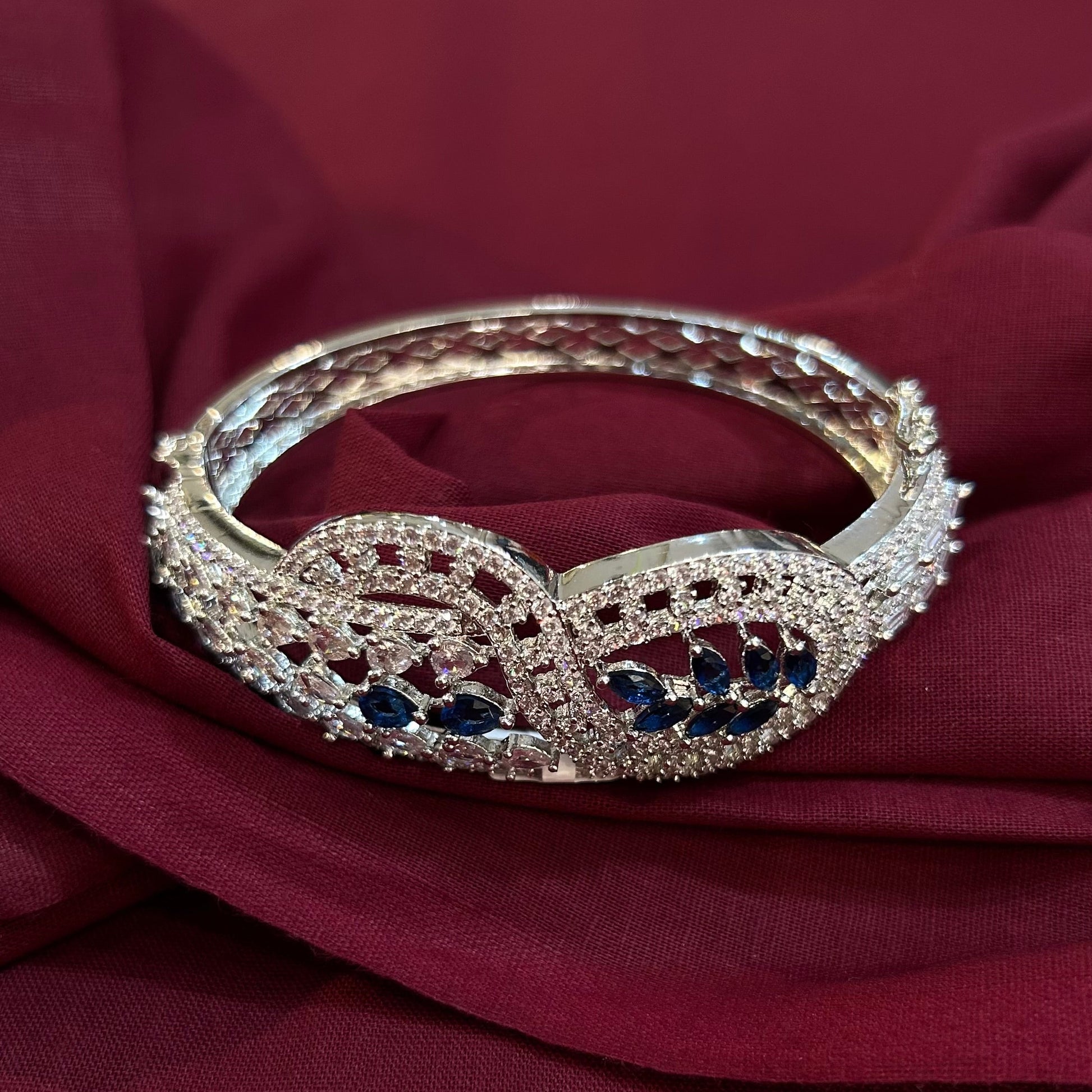 A silver-toned filigree bangle bracelet with dark blue gemstone accents, displayed on draped burgundy fabric.