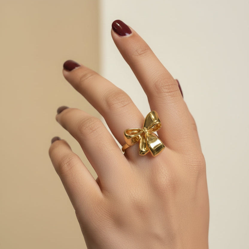 A gold ring with a bow and small hanging charms, worn on a person's ring finger, shown against a dark textured background.