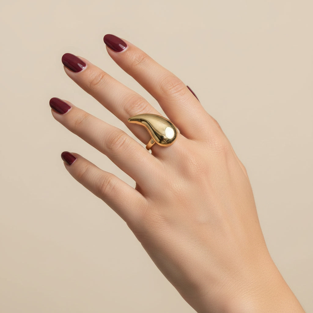 A gold ring with a teardrop-shaped setting worn on a person's ring finger, shown against a black textured leather background.