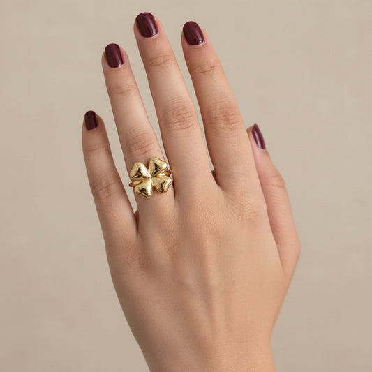 A close-up of a hand wearing a large, textured gold ring with a floral or clover-like design, resting on a dark, curved surface.