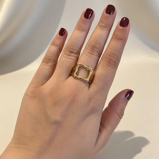 A gold ring with a square openwork design worn on a person's ring finger, shown against a dark textured surface.