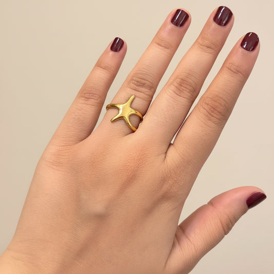 A gold ring shaped like a five-pointed star or flower, worn on the middle finger of a hand resting on a black textured surface.